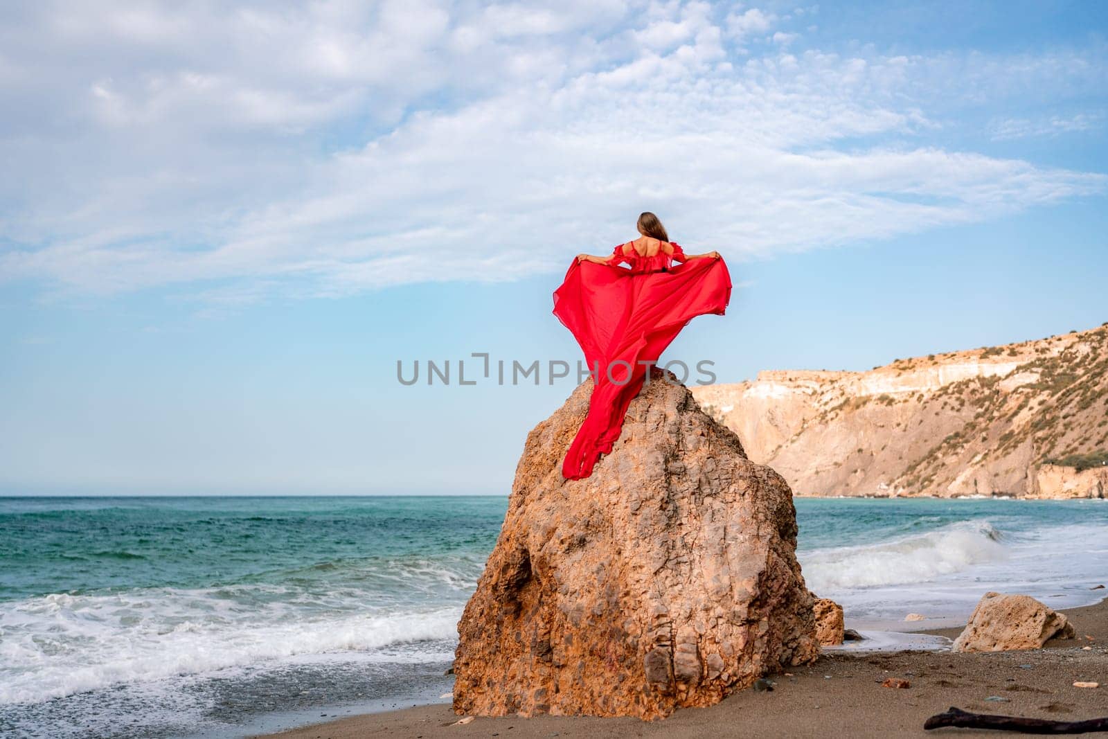 woman sea red dress. Woman with long hair on a sunny seashore in a red flowing dress, back view, silk fabric waving in the wind. Against the backdrop of the blue sky and mountains on the seashore. by Matiunina