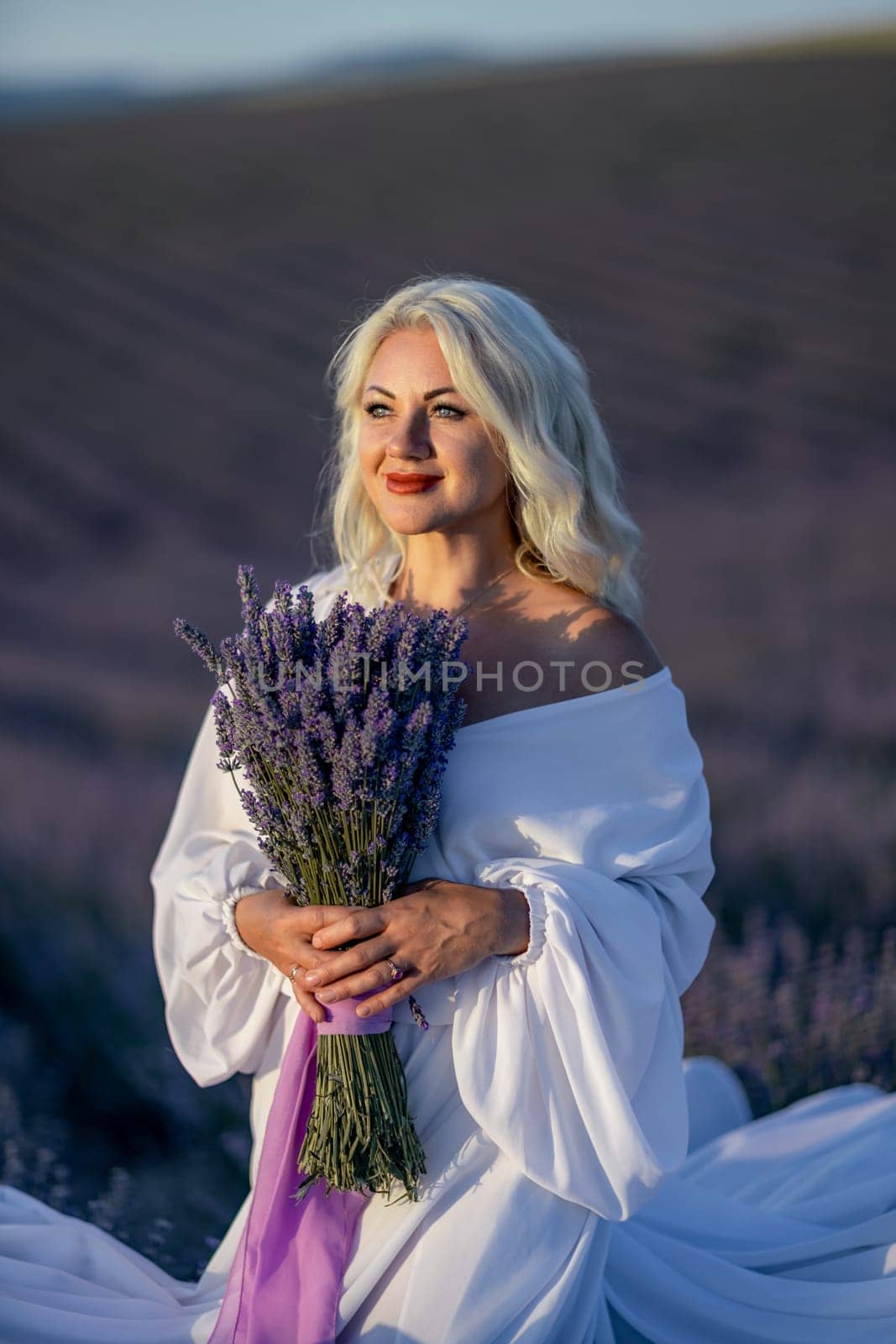 Blonde woman poses in lavender field at sunset. Happy woman in white dress holds lavender bouquet. Aromatherapy concept, lavender oil, photo session in lavender.