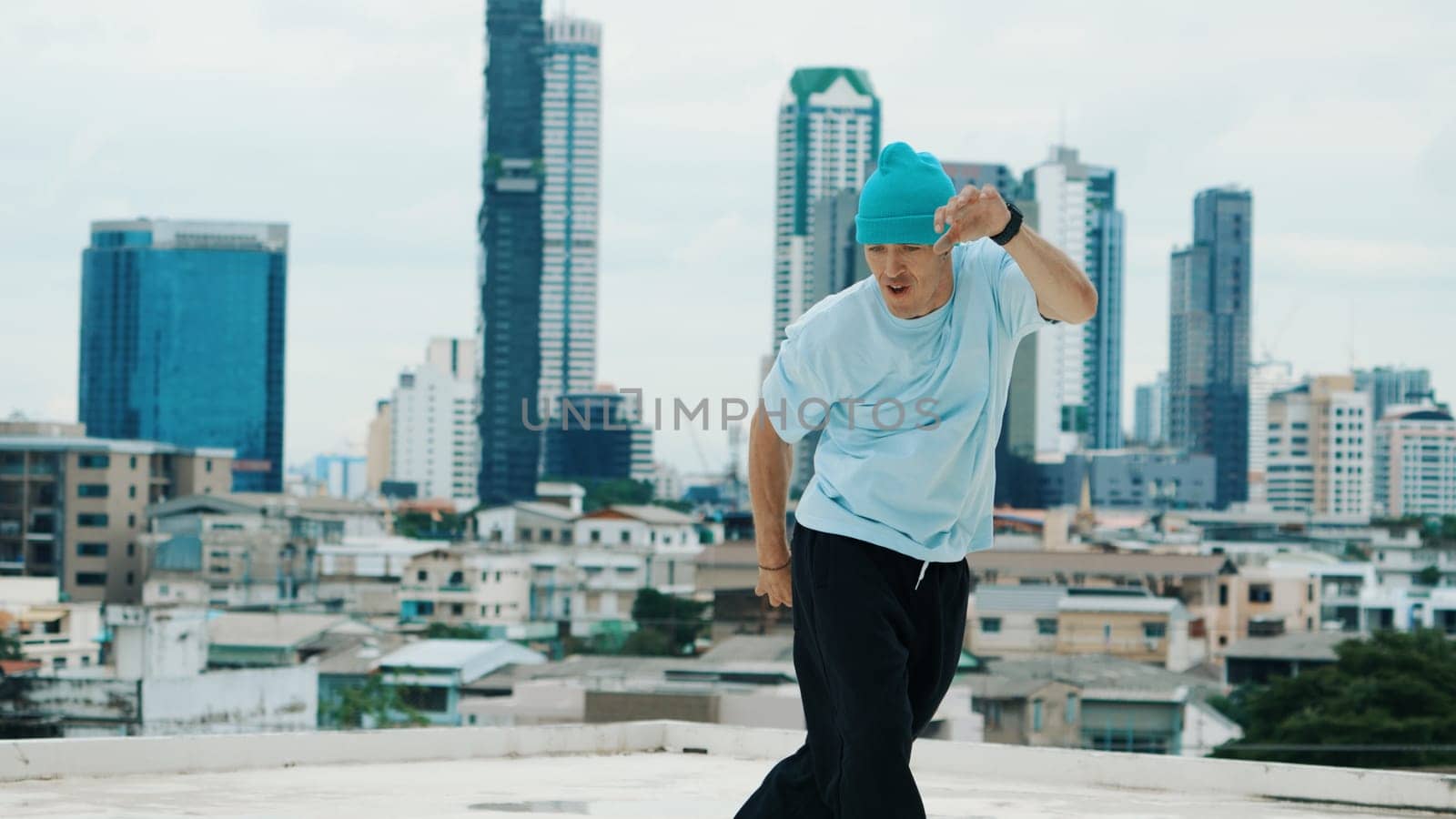 Caucasian B-boy dancer practicing street dancing at rooftop with city or urban. Motion shot of young man performing street dance by doing freeze pose. Break dance. Outdoor sport 2024. Endeavor.