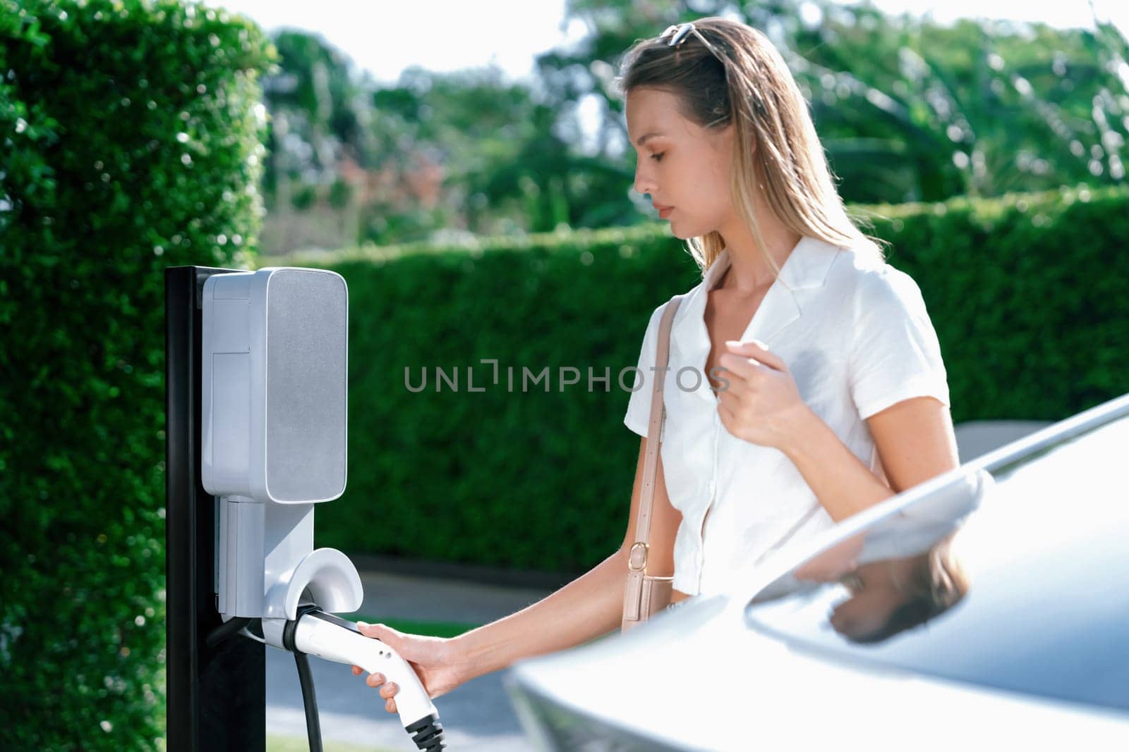 Young woman travel with EV electric car charging in green sustainable city outdoor garden in summer. Urban sustainability lifestyle by green clean rechargeable energy of electric BEV vehicle innards