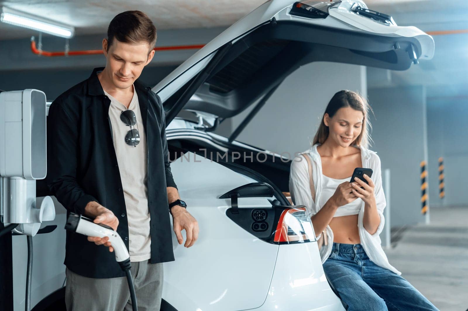 Young couple travel with EV electric car to shopping center parking lot charging in downtown city showing urban sustainability lifestyle by green clean rechargeable energy of electric vehicle innards