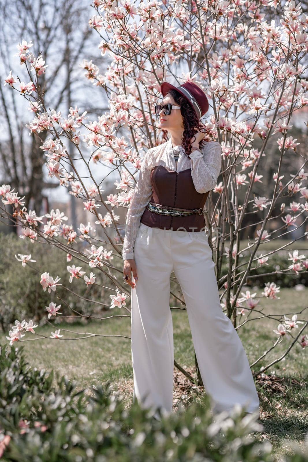 Magnolia park woman. Stylish woman in a hat stands near the magnolia bush in the park. Dressed in white corset pants and posing for the camera. by Matiunina