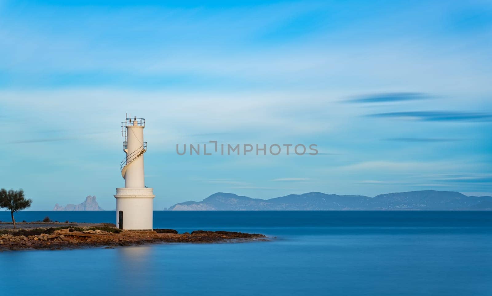 Photo captures long exposure of a lighthouse on Formentera's coast with silhouetted Es Vedra, Ibiza island in the backdrop.