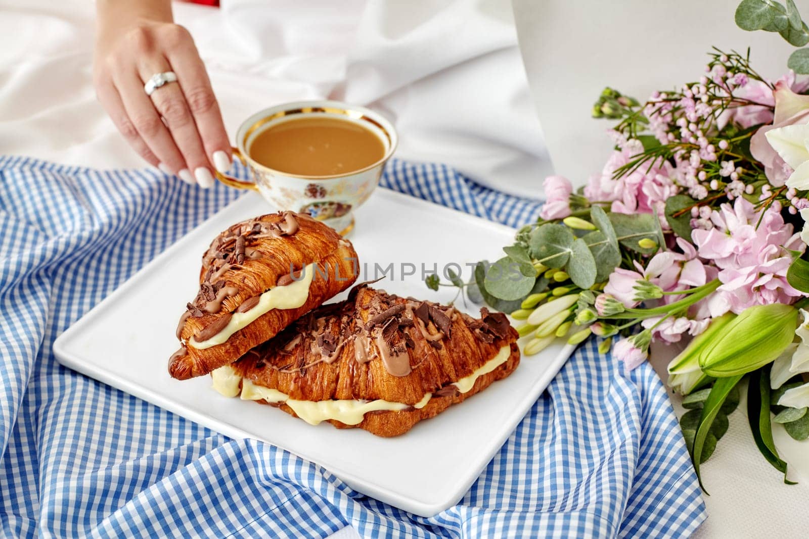 Romantic intimate breakfast in bed scene with woman reaching for cup of coffee, fresh French croissants with delicate custard and chocolate, and lush flower bouquet on white sheets, cropped shot