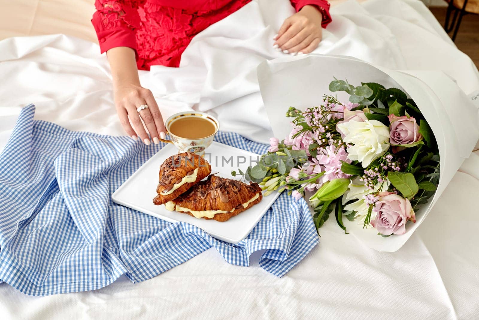 Elegant romantic morning setting with woman in red enjoying breakfast in bed with fresh French chocolate croissants filled with delicate custard and tender flower bouquet on white sheets, cropped shot