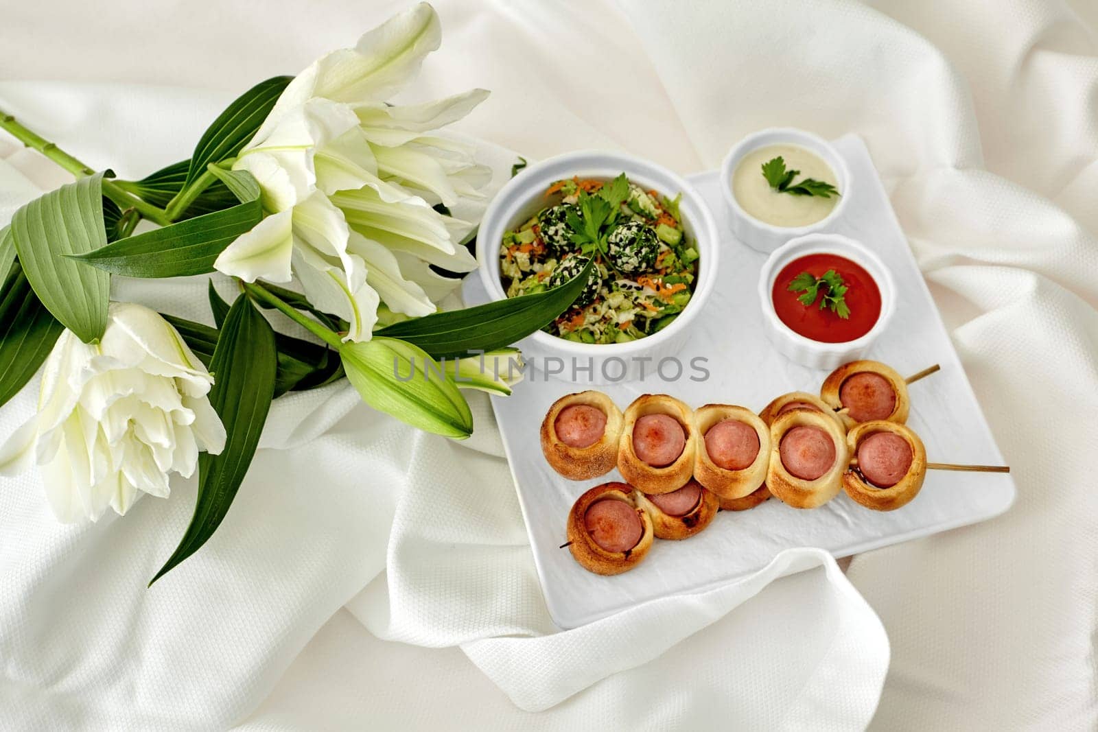 Elegant romantic snack arrangement featuring pigs in blankets on skewers, fresh green salad, dipping sauces, and fragrant white lilies on white fabric background