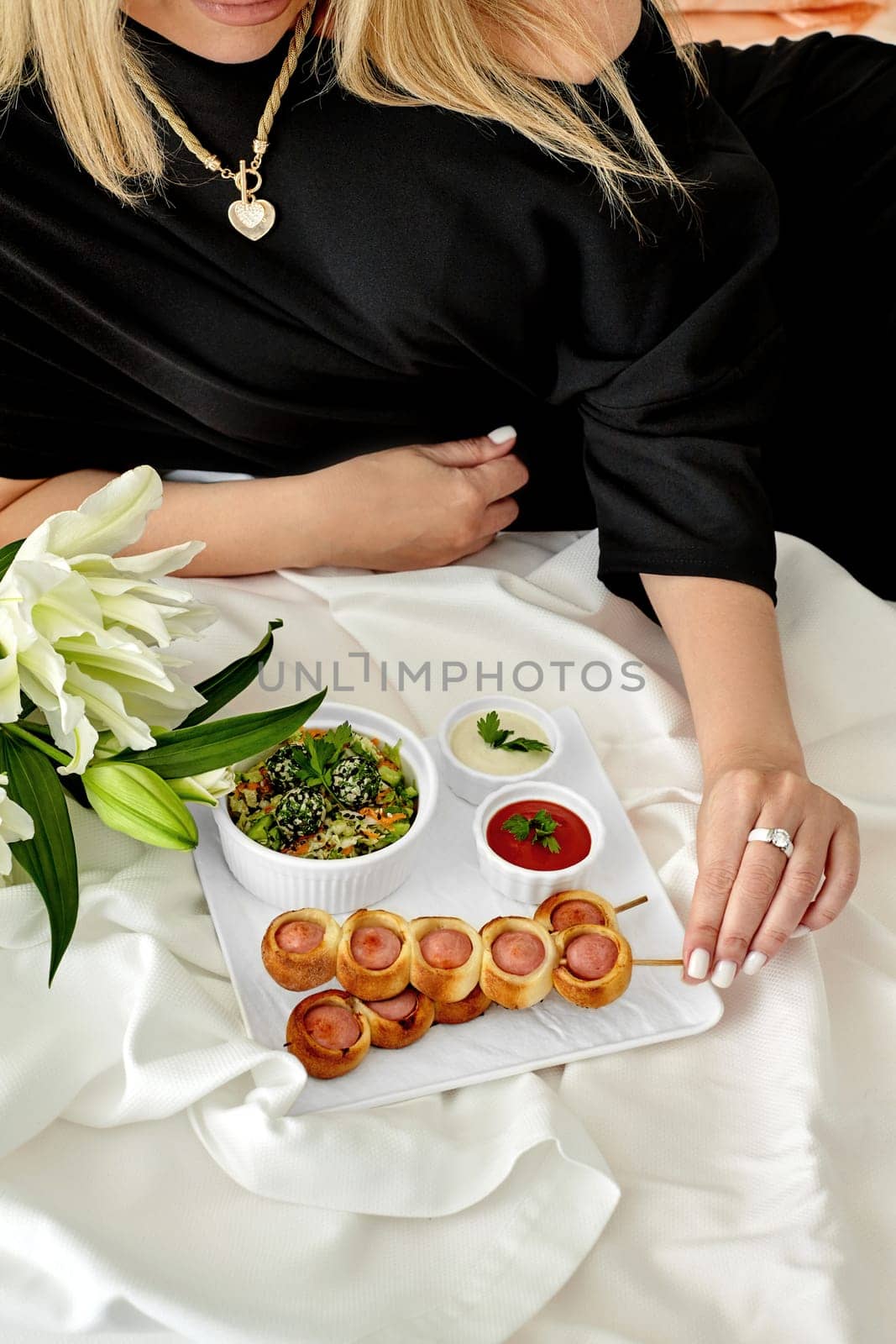 Sophisticated woman relaxing on white bedding with tray of delicious freshly baked sausage rolls, healthy vegetable salad, spicy dipping sauces, and fragrant white lilies. Romantic weekend concept