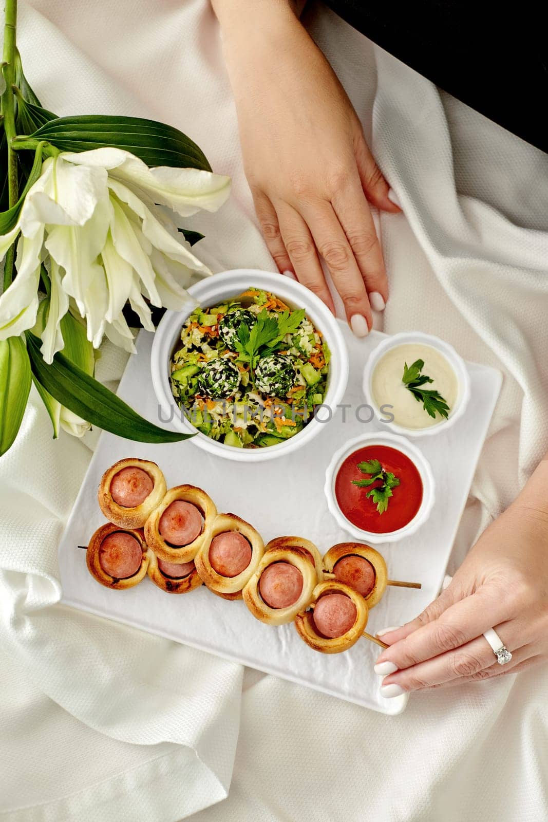 Female hand taking appetizing golden brown sausage rolls on skewers served on plate with vegetable salad and sauces against white tablecloth background decorated with fresh lilies