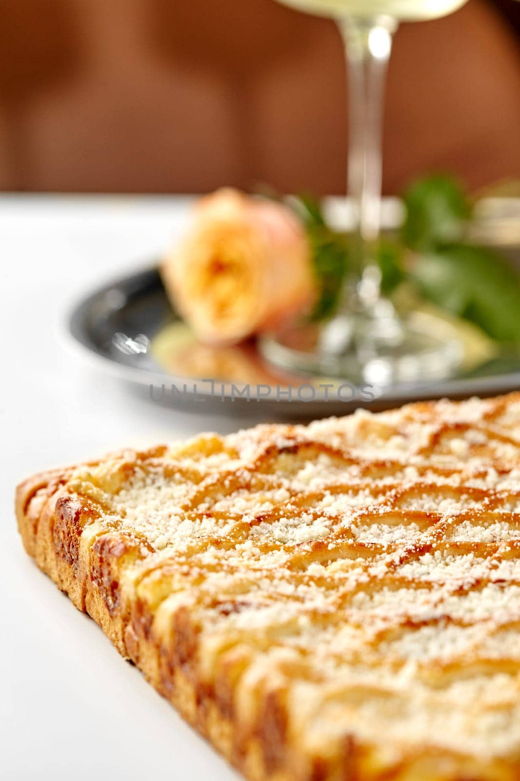 Selective focus on delicious golden lattice cheese galette topped with parmesan crumbs served on table in romantic setting with glass of wine and fresh rose on blurred background
