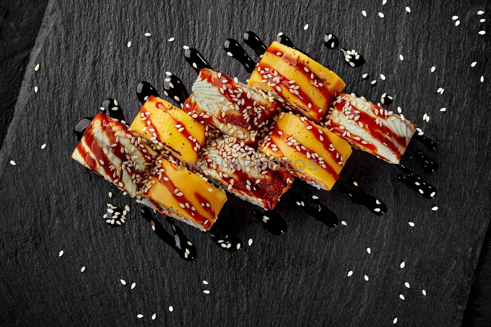 Top view of uramaki rolls wrapped in cheddar slices and eel fillets dressed with tangy unagi sauce and sprinkled with sesame seeds on black slate board. Traditional Japanese cuisine