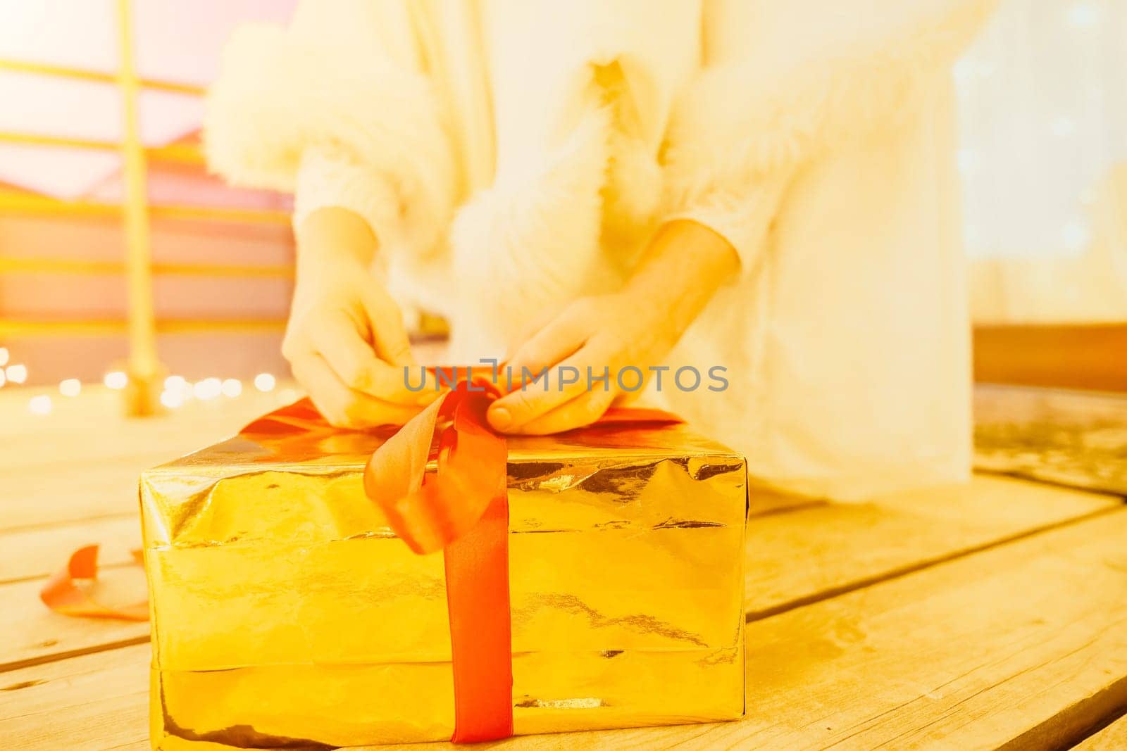 A woman in a white dress is holding a gold box with a red ribbon. She is wearing a crown on her head. The scene takes place in a room with a door and a window. The woman appears to be opening the gift box.