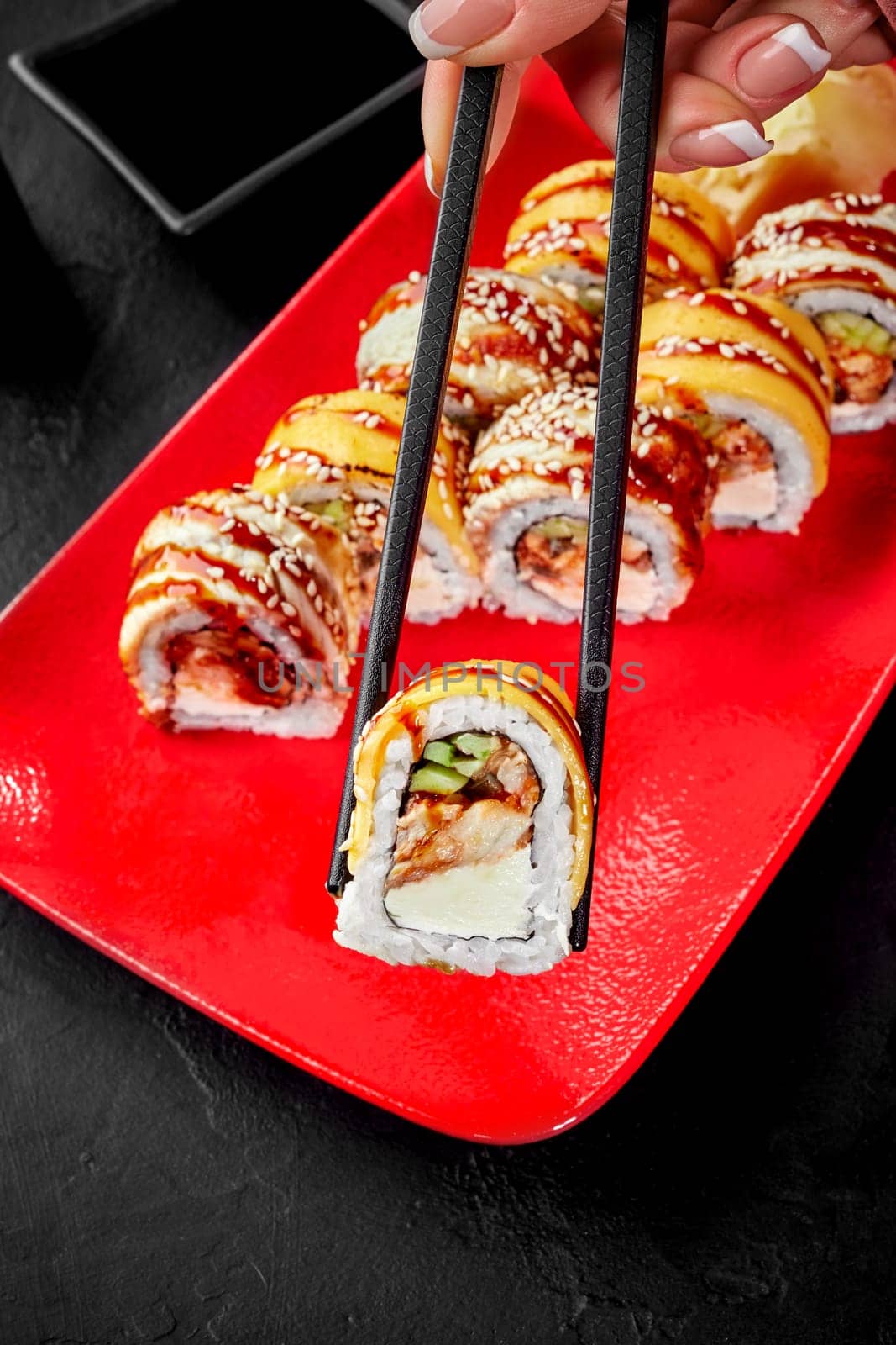 Close-up of female hand using chopsticks to pick up appetizing sushi roll filled with eel, cucumber and cream cheese topped with cheddar from vibrant red plate against black background