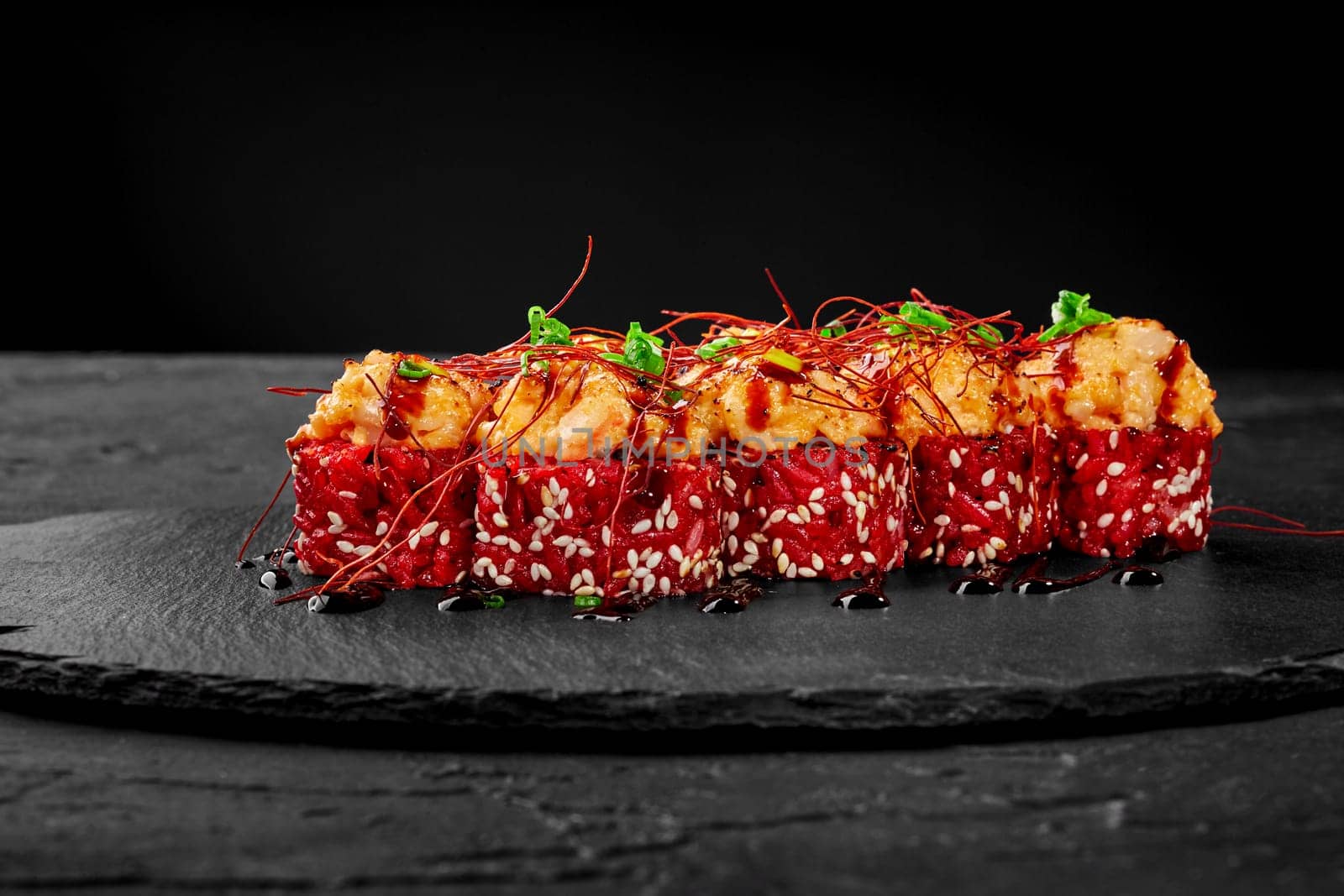 Closeup of appetizing red rice sushi rolls topped with scallop cheese caps seasoned with tangy unagi sauce garnished with chopped scallion served on black slate board. Japanese cuisine