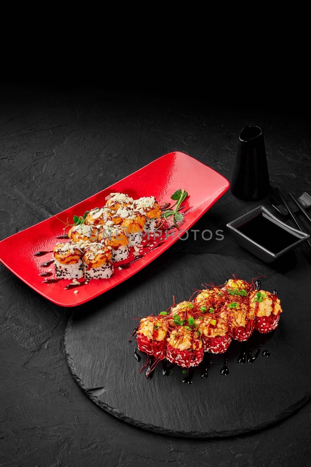 Elegant presentation of two kinds of sushi rolls with seared cheese and seafood caps served of on striking red plate and black slate, traditionally accompanied by soy sauce and chopsticks