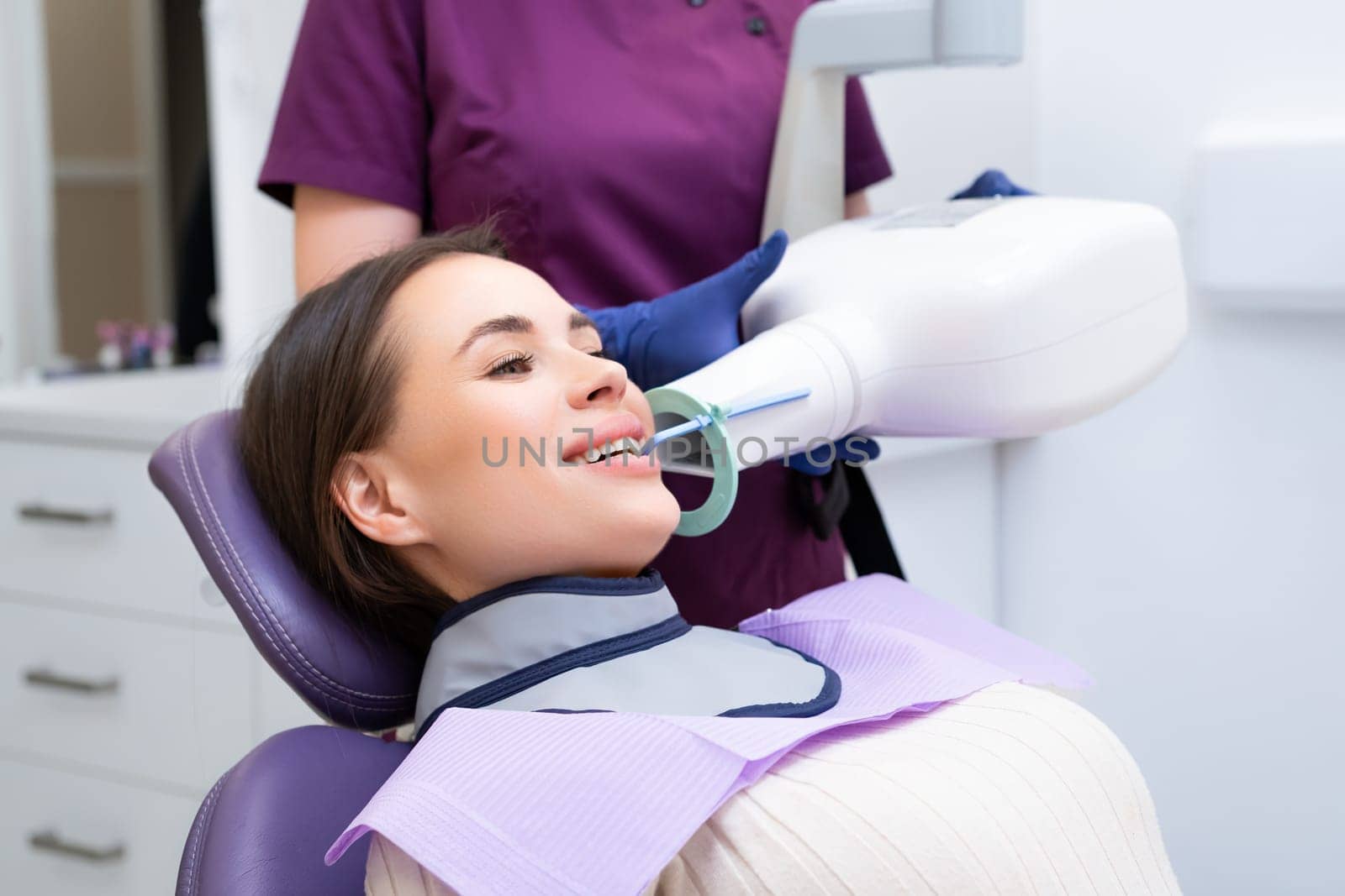 Dental assistant prepares patient for X-ray using specialized tools in clinic. Woman asks female patient to smile for better view of mouth