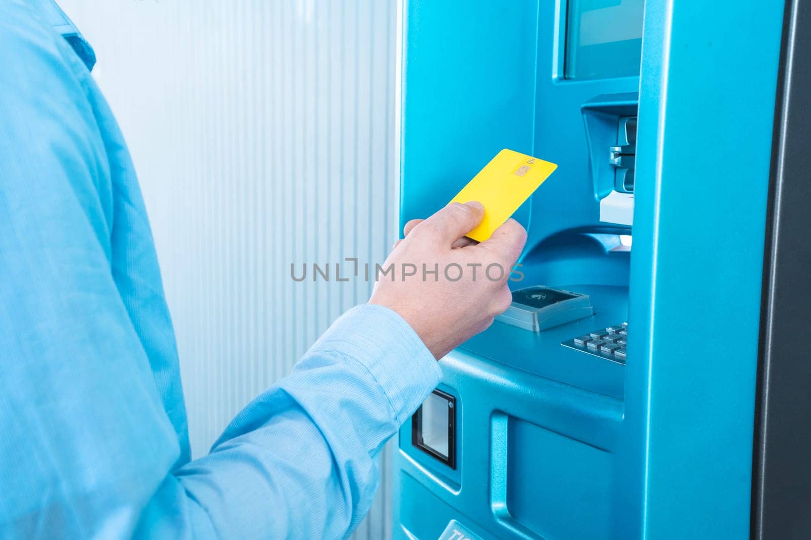 A man is inserting a blue credit card into a plastic ATM machine in a room filled with electric blue paint and fluid plastic bottles