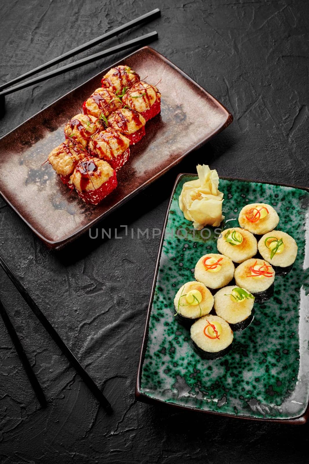 Two kinds of appetizing baked sushi rolls with soft golden cheese hats garnished with spicy mayo, vegetable shavings and chili threads served on colorful ceramic plates on black background