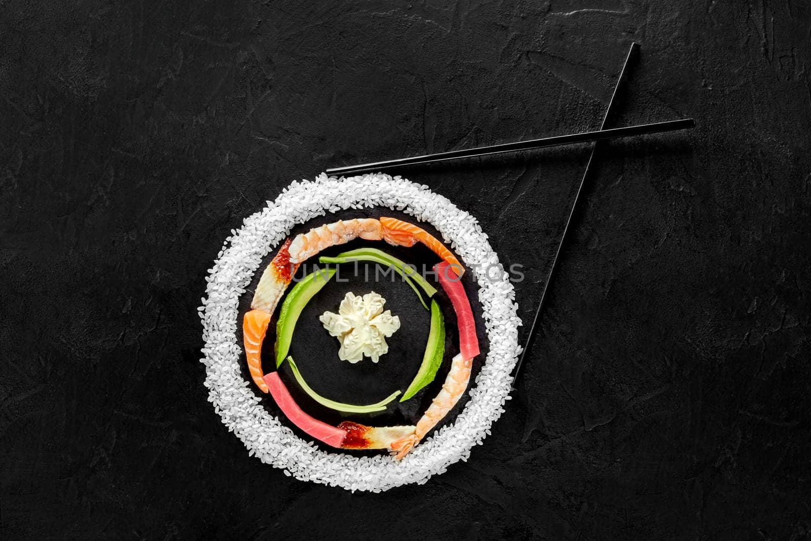 Layered colorful circle of raw rice, salmon fillet, eel, tuna and shrimps, avocado slices and cream cheese with chopsticks laid out on black background. Traditional Japanese sushi ingredients