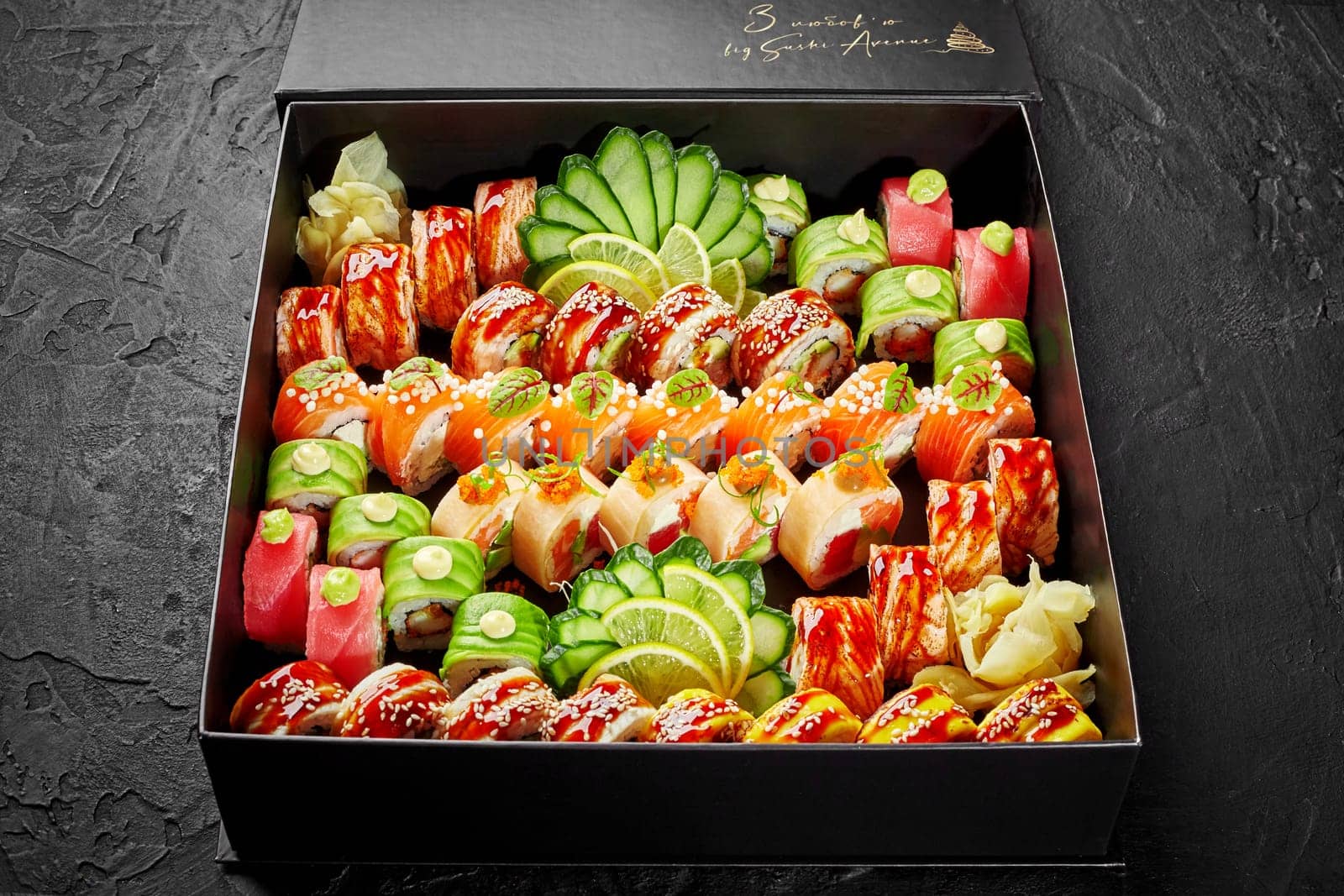 Large set of various Japanese rolls with tuna, salmon and eel packed in cardboard box with pickled ginger, slices of cucumbers and lemon, on black background. Sushi delivery concept