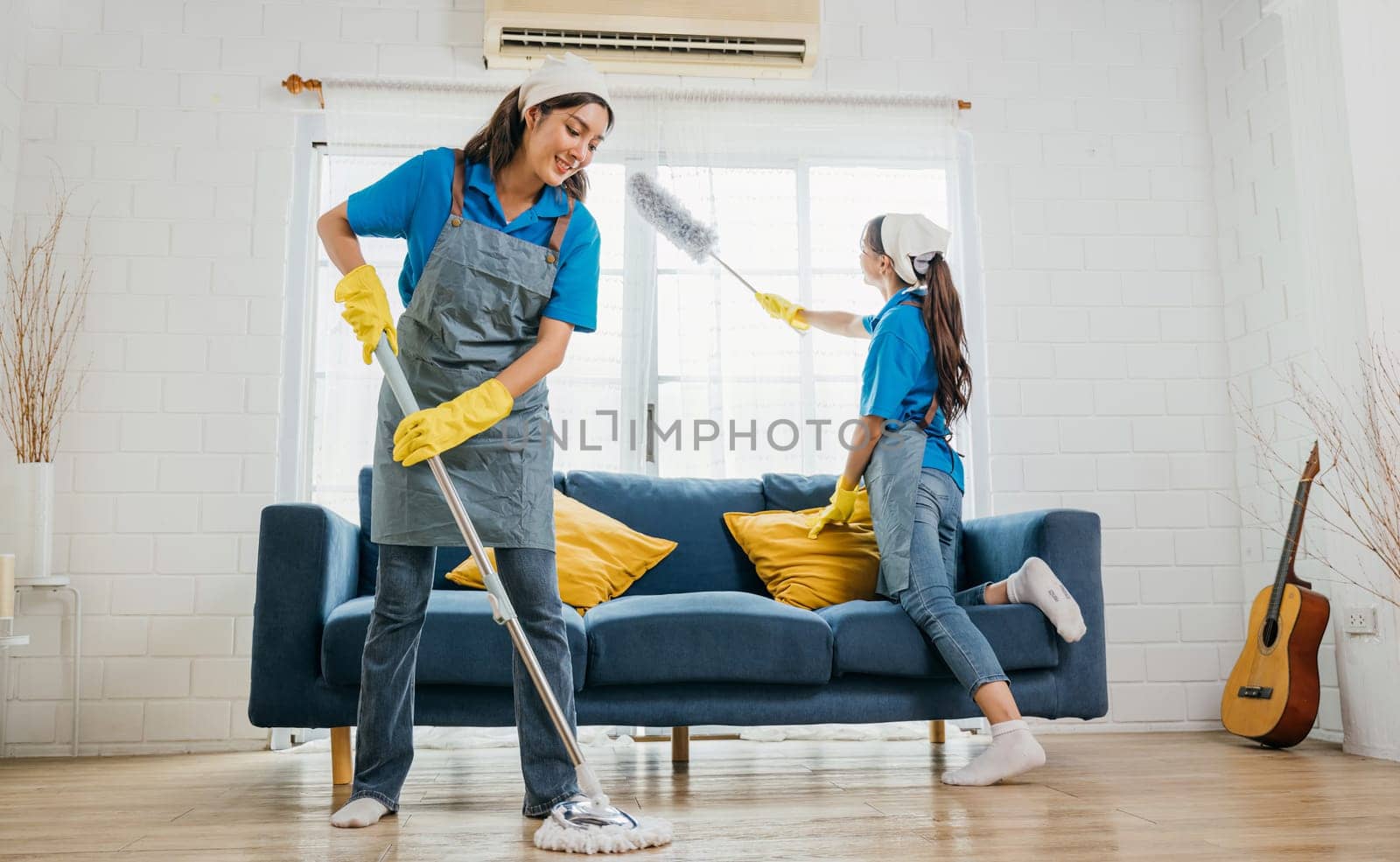 Women in gloves and uniforms employees of a cleaning service carefully clean a dusty living room. They're holding sprays and mops ensuring purity and hygiene. House care is their occupation. by Sorapop