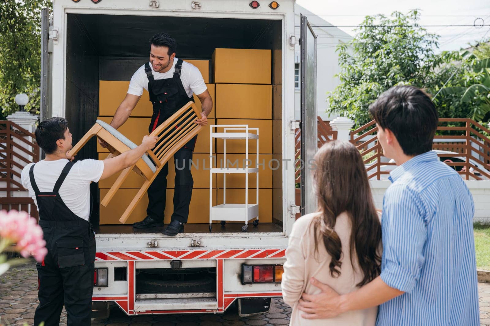 Assisted by a professional delivery team a couple moves into their new house. Employees work together unloading and lifting cardboard boxes for a smooth relocation. Moving Day Concept by Sorapop