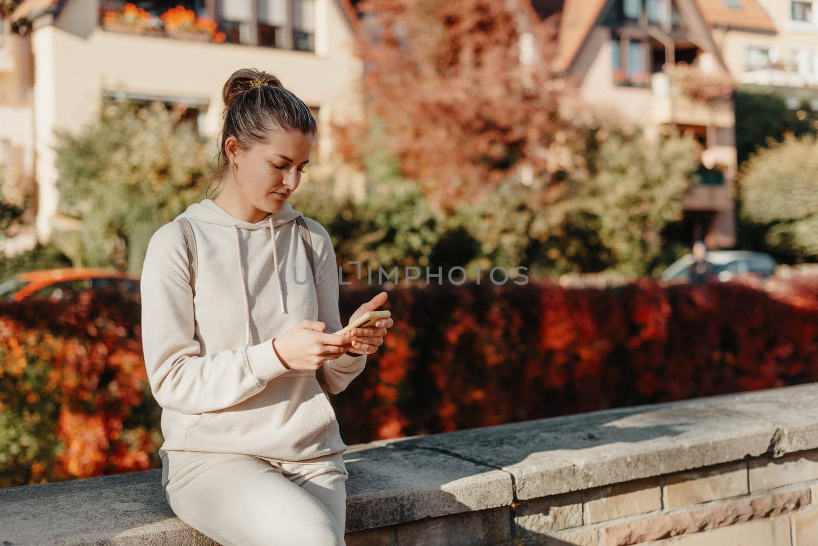 Young Fashionable Teenage Girl With Smartphone In Europian Park In Autumn Sitting At Smiling. Trendy Young Woman In Fall In Park Texting. Retouched, Vibrant Colors. Beautiful Blonde Teenage Girl Wearing Casual Modern Autumn Outfit Sitting In Park In Autumn. Retouched, Vibrant Colors, Brownish Tones. by Andrii_Ko