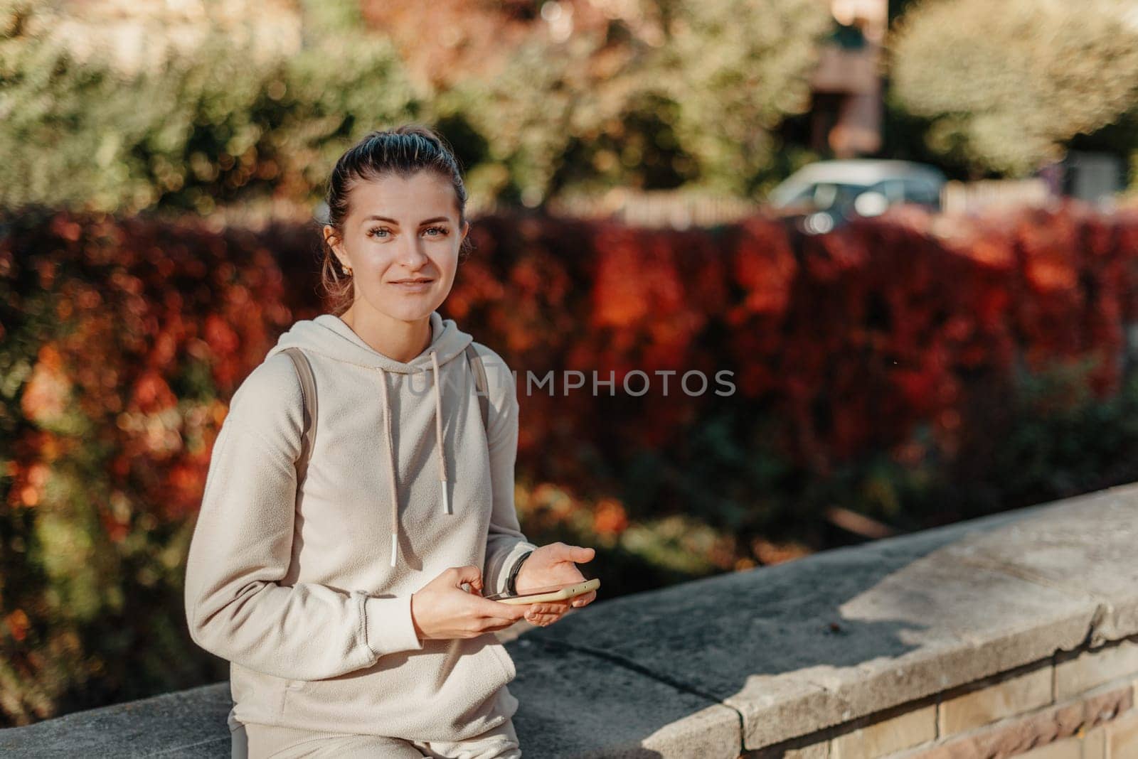 Young Fashionable Teenage Girl With Smartphone In Europian Park In Autumn Sitting At Smiling. Trendy Young Woman In Fall In Park Texting. Retouched, Vibrant Colors. Beautiful Blonde Teenage Girl Wearing Casual Modern Autumn Outfit Sitting In Park In Autumn. Retouched, Vibrant Colors, Brownish Tones. by Andrii_Ko