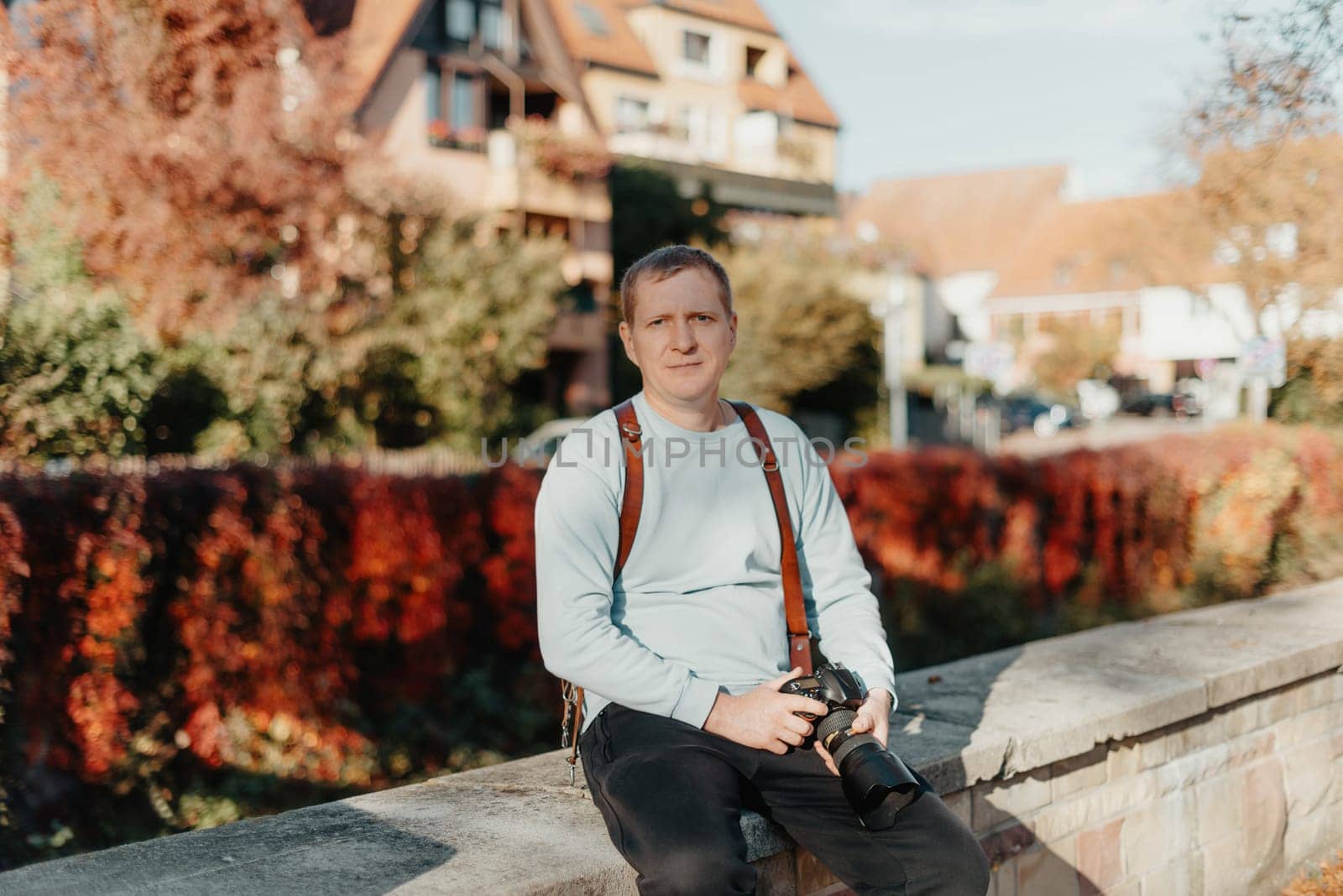 Man Sitting in Old European City And Holding Photo Camera. Contemporary Stylish Blogger And Photographer. Handsome man taking a selfie on a trip in Europe. by Andrii_Ko