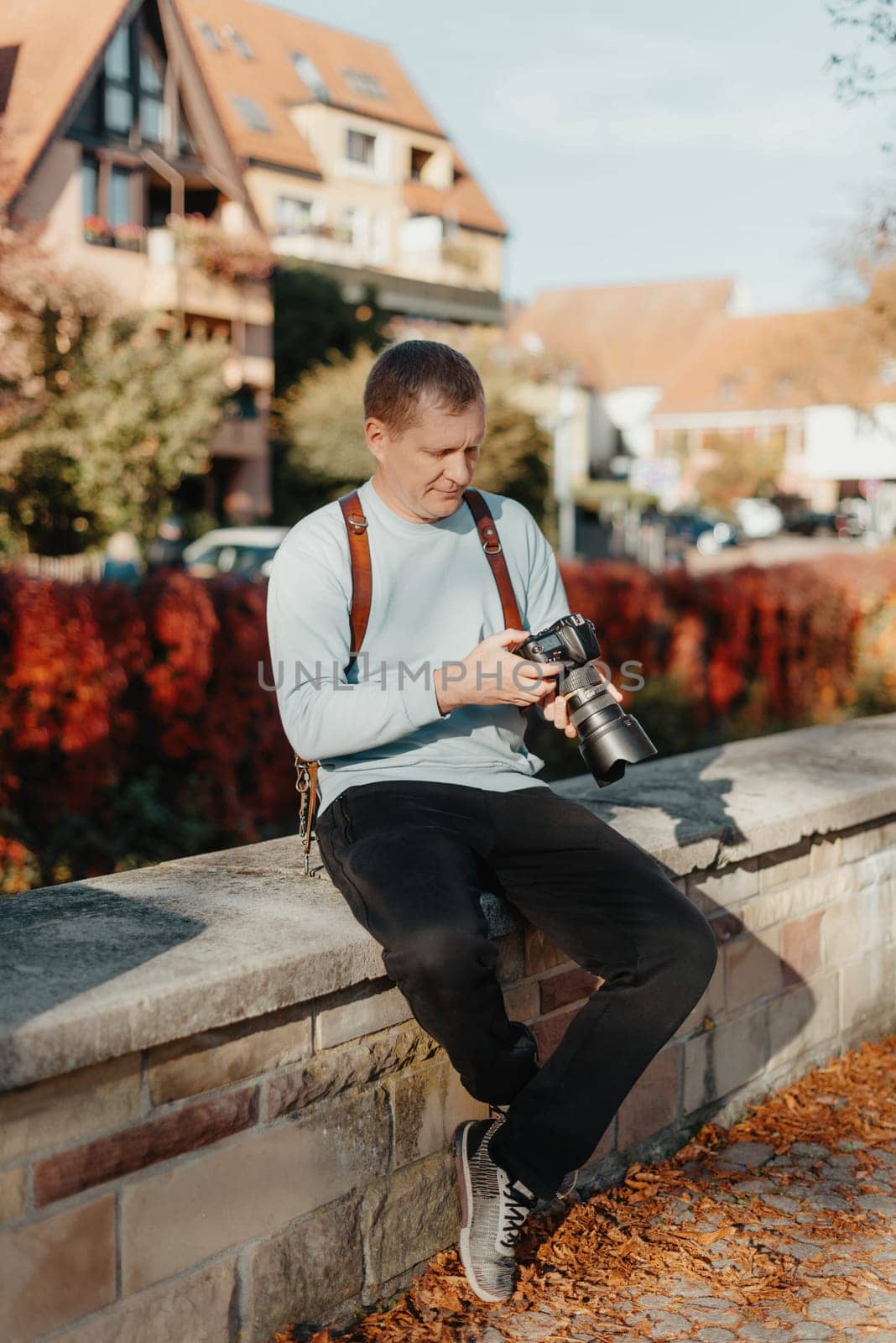 Man Sitting in Old European City And Holding Photo Camera. Contemporary Stylish Blogger And Photographer. Handsome man taking a selfie on a trip in Europe. by Andrii_Ko