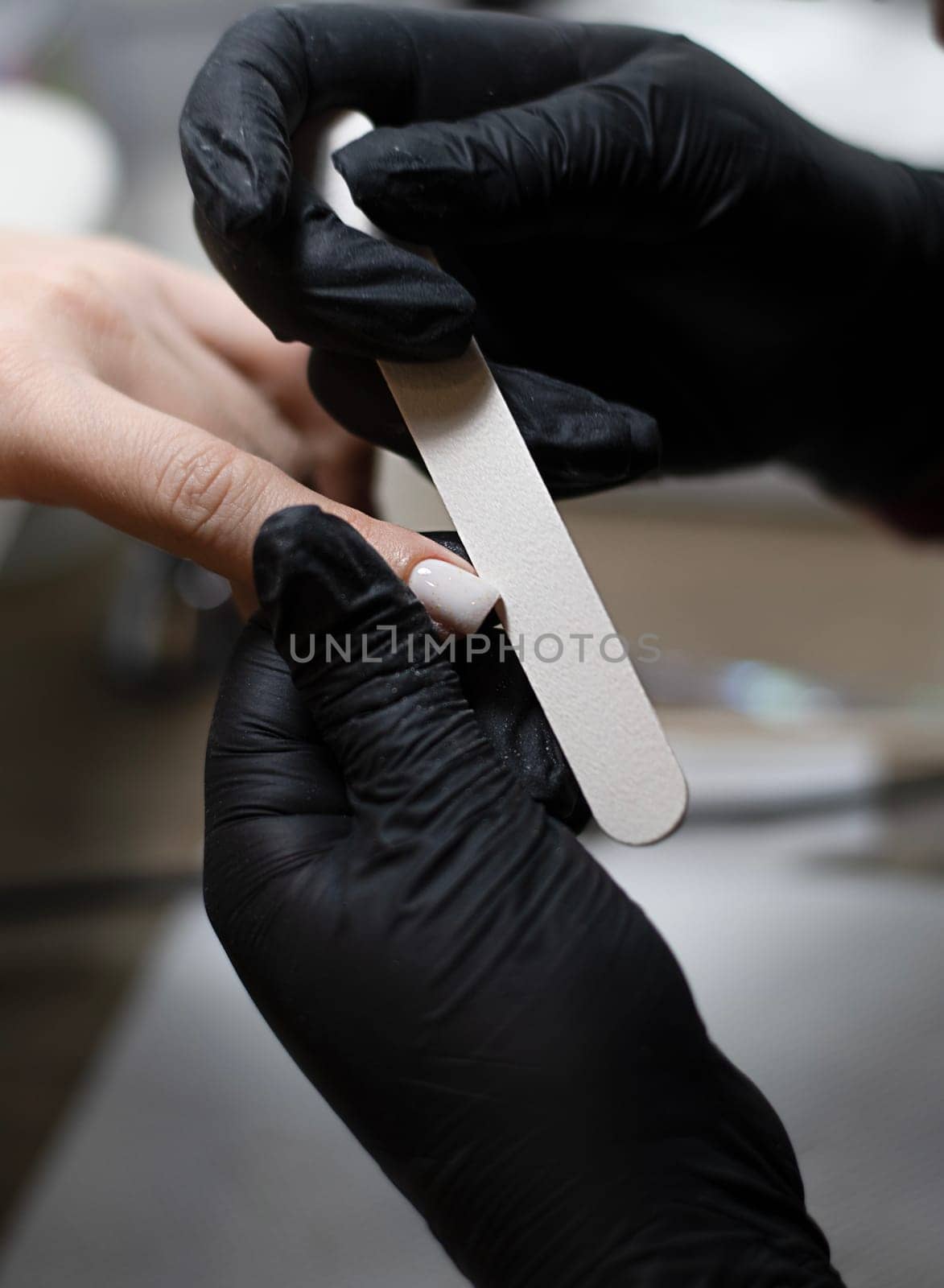 Beauty concept. A manicurist in black latex gloves makes a hygienic manicure, paints the client's nails with gel polish and files them with a nail file in a beauty salon. Close-up. Vertical.