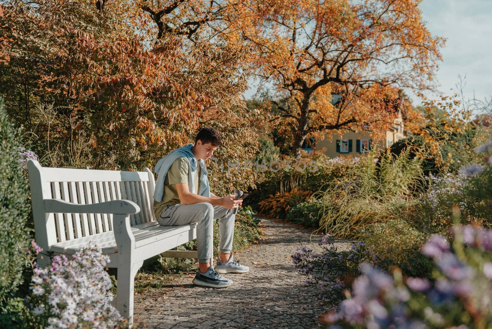 A Teenager Sits On A Bench In The Autumn Park Drinks Coffee From A Thermo Mug And Looks Into A Phone. Portrait Of Handsome Cheerful Guy Sitting On Bench Fresh Air Using Device Browsing Media Smm Drinking Latte Urban Outside Outdoor by Andrii_Ko