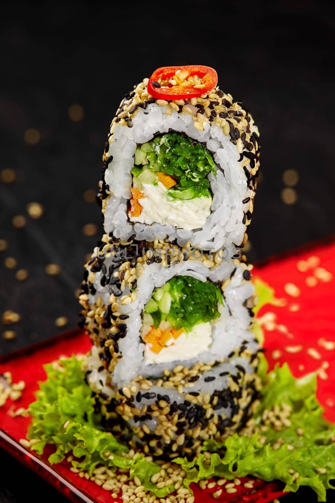 Closeup of appetizing light vegetarian sesame sushi rolls with juicy filling of cream cheese, carrot, hiyashi wakame, lettuce and cucumber garnished with spicy chili pepper slice, stacked on red plate