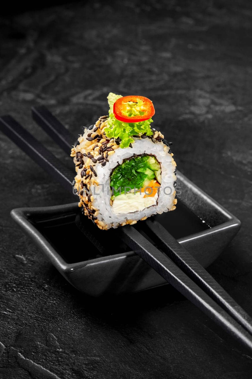 Vegetarian sushi roll with light filling of cream cheese, carrot, hiyashi wakame, lettuce and cucumber adorned with sesame seeds and chili, perfectly balanced on chopsticks over soy sauce dish