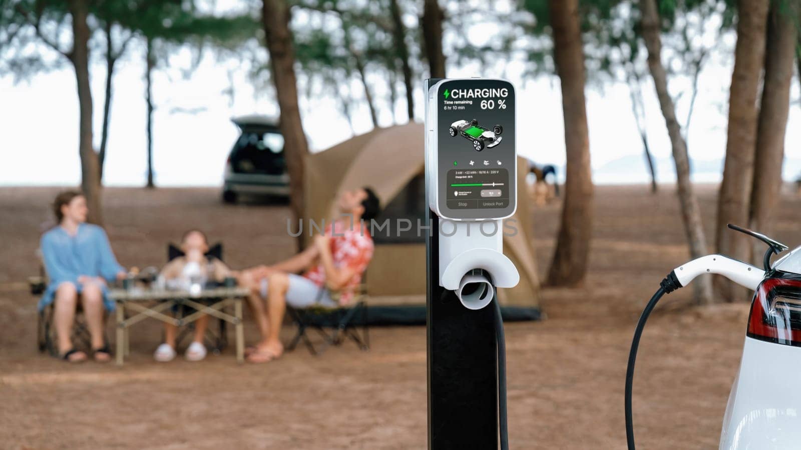 Alternative family camping trip traveling by the beach with electric car recharging battery from EV charging station with blurred family enjoying the seascape campsite background. Perpetual