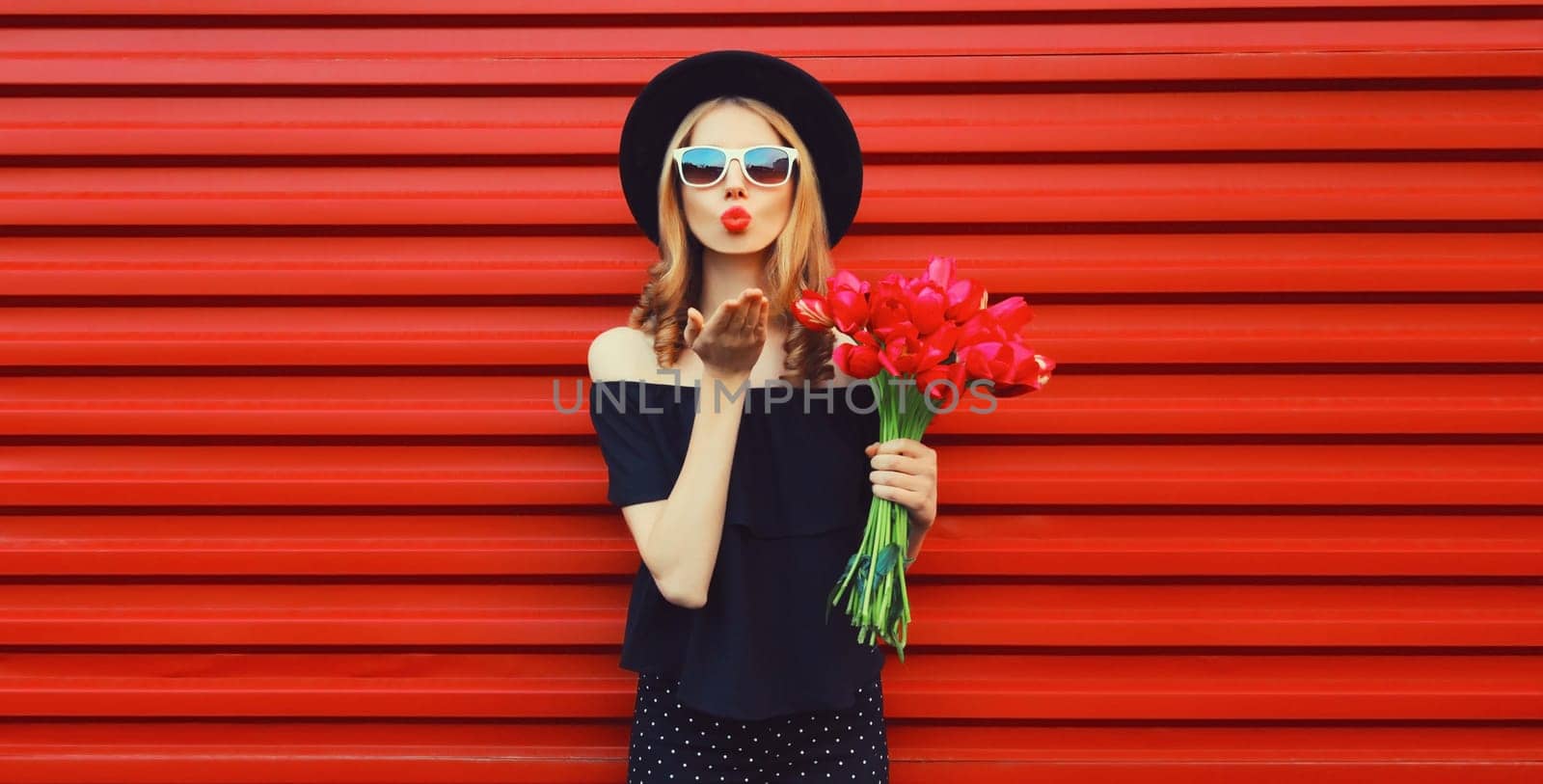 Portrait of beautiful stylish woman with bouquet of red rose flowers blowing kiss in black round hat