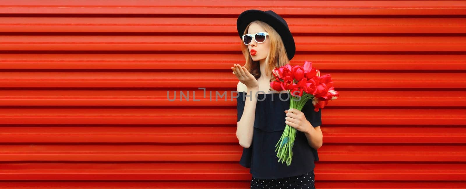 Portrait of beautiful stylish woman with bouquet of red rose flowers blowing kiss in black round hat