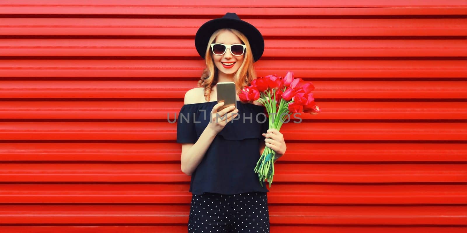 Beautiful smiling woman with smartphone holding bouquet of red rose flowers in black round hat