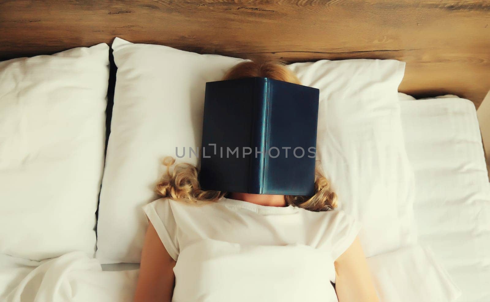 Young sad tired woman student covering her face with open book with blank cover while lying on the bed at home
