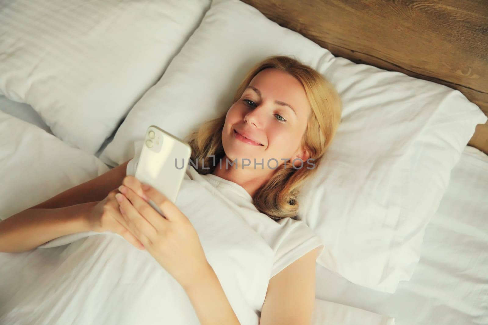 Healthy sleep, happy relaxed young woman with mobile phone lying in bed on white soft comfortable pillow at home