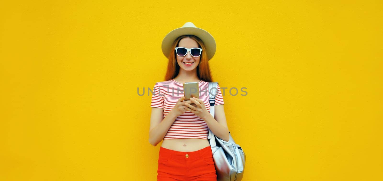 Summer portrait of happy traveler young woman 20s with mobile phone looking at device in casual straw hat with backpack on bright yellow studio background