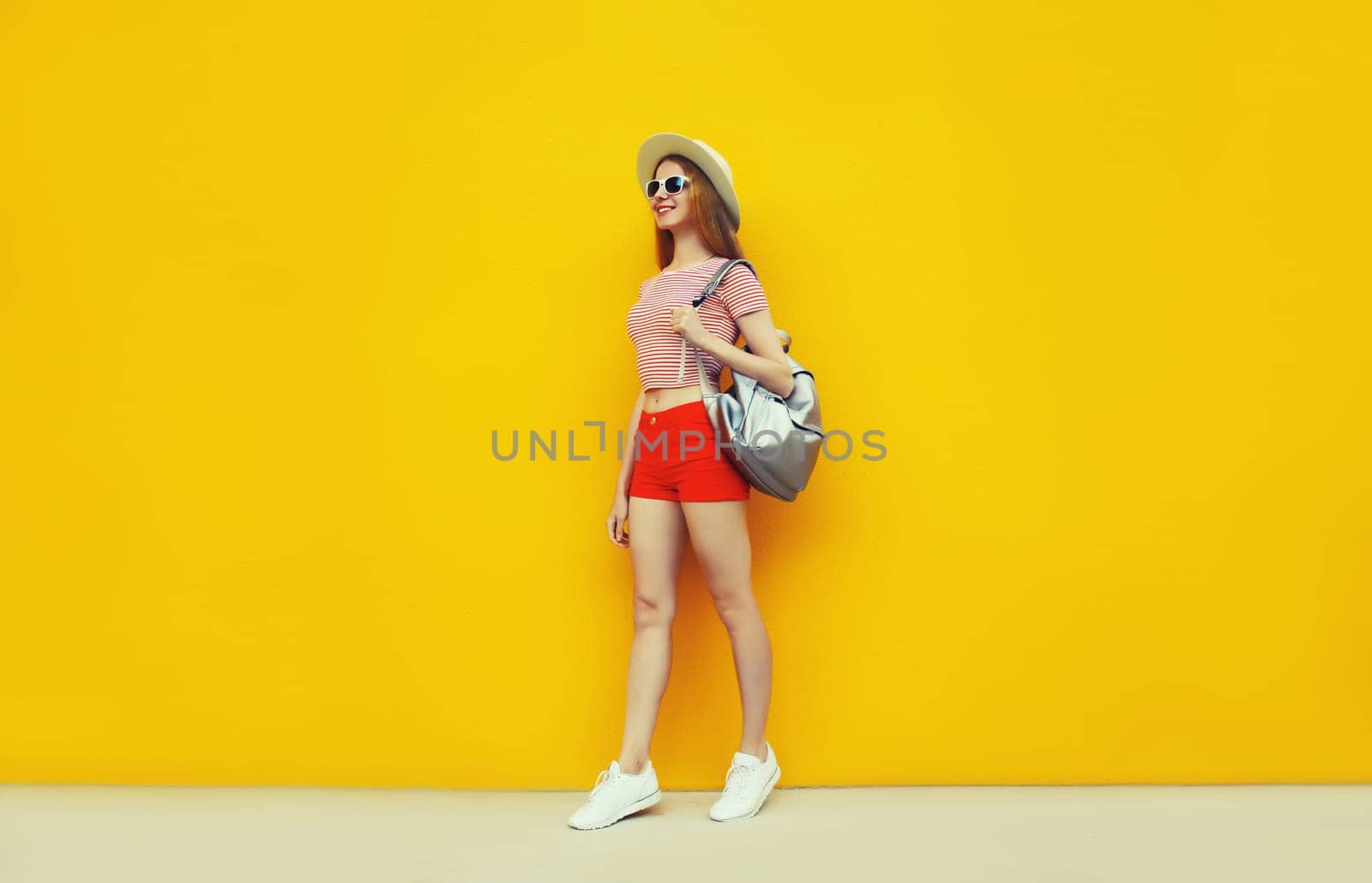 Beautiful young woman full length standing in summer straw hat, red shorts with backpack on vivid yellow background