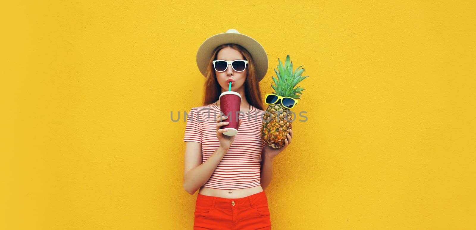 Summer bright portrait of stylish young woman drinking fresh juice holding pineapple fruit in straw hat, sunglasses on colorful yellow studio background