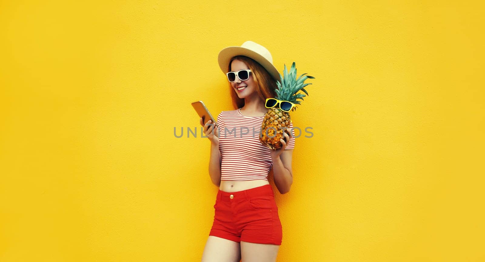 Summer portrait of traveler happy young woman with mobile phone holding fresh pineapple fruit looking at device in casual straw hat, sunglasses on bright yellow studio background