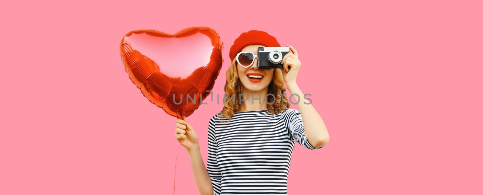 Summer portrait of happy smiling young woman with film camera and red heart shaped balloon wearing french beret hat on pink studio background