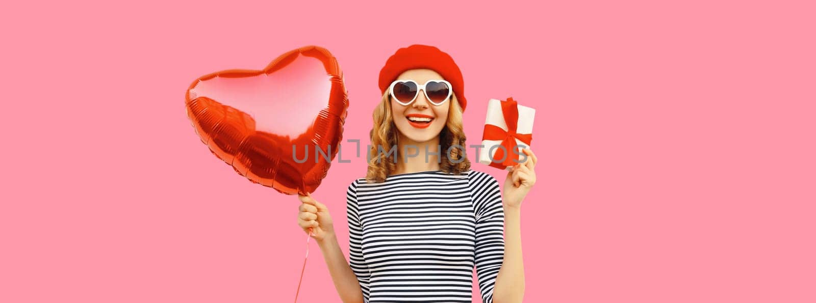 Cute portrait of happy smiling young woman with red heart shaped balloon and gift box wearing french beret hat on pink studio background