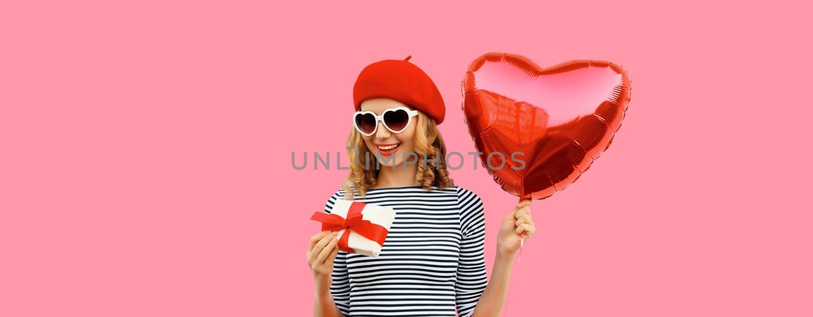 Cute portrait of happy smiling young woman with red heart shaped balloon and gift box wearing french beret hat on pink studio background