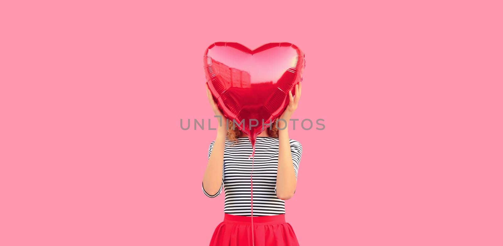 Cute portrait of happy sweet woman covering her head with red heart shaped balloon on pink studio background