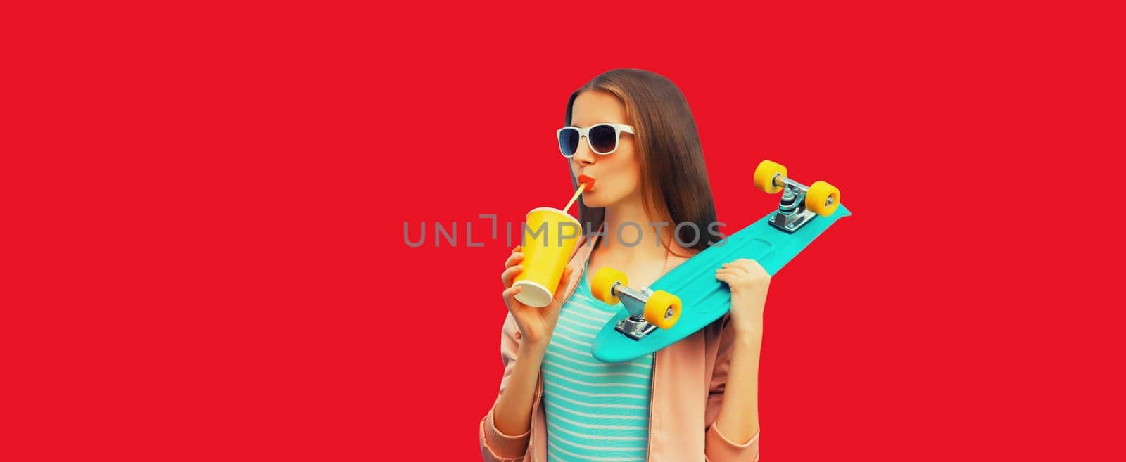 Portrait of stylish young woman with skateboard drinking juice on red background