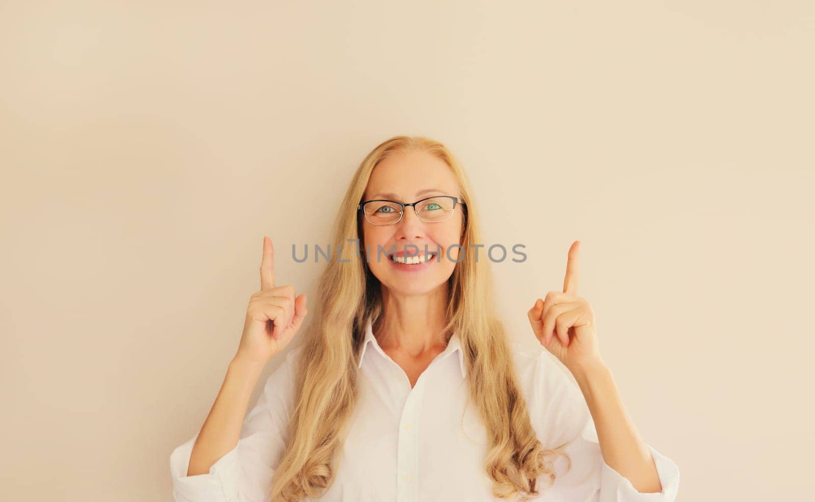 Happy exciting smiling middle aged woman in eyeglasses pointing her fingers up and looking at camera