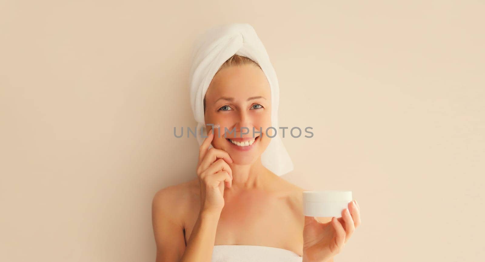 Natural beauty portrait of happy middle-aged woman touches her clean skin applying face cream with bath towel on head after shower in morning